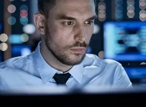 Man in blue shirt works at computer in server room, focused expression.