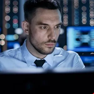 Man in blue shirt works at computer in server room, focused expression.