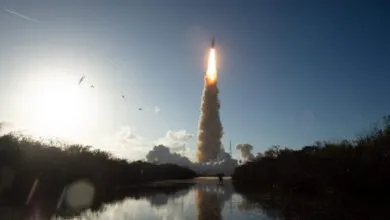 Rocket launch with bright flames and smoke rising into a blue sky, reflected in water.