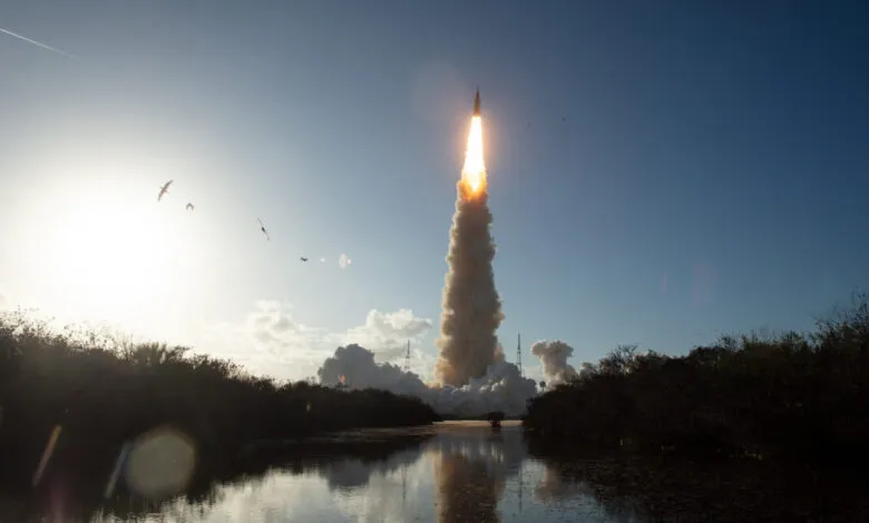 Rocket launch with bright flames and smoke rising into a blue sky, reflected in water.