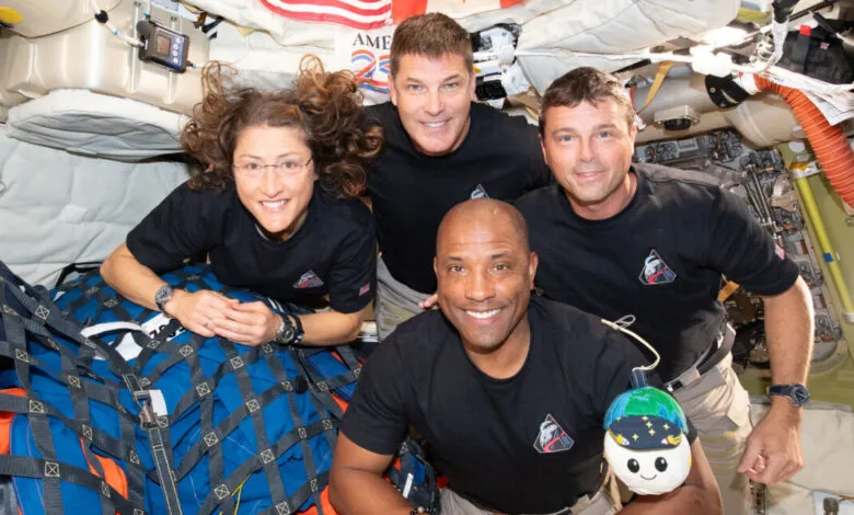 Four astronauts smile in the International Space Station wearing matching shirts.