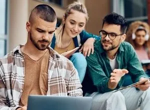Three college students collaborate on a laptop outdoors on campus steps.