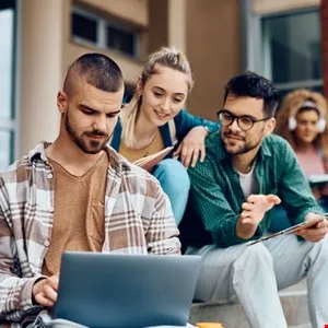 Three college students collaborate on a laptop outdoors on campus steps.