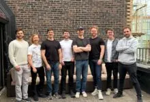 Eight men stand together outdoors against a brick wall and spiral staircase.