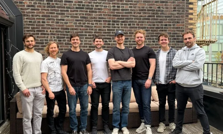 Eight men stand together outdoors against a brick wall and spiral staircase.