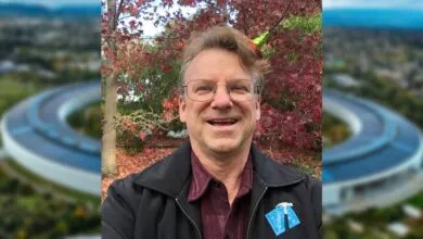 Man smiles in front of Apple Park, wearing glasses and a black jacket.