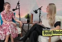 Two women sit in chairs for a podcast recording, smiling at each other.