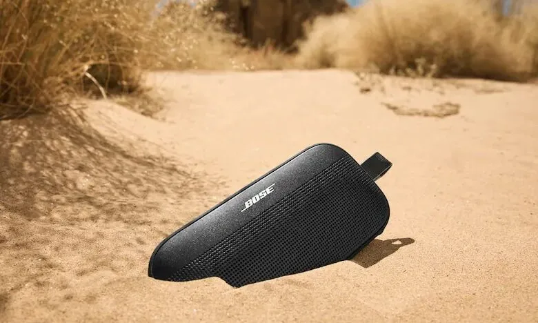 Black Bose SoundLink Flex speaker on sand with desert plants in background.