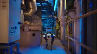 Robot dog walks through industrial setting with pipes and equipment.