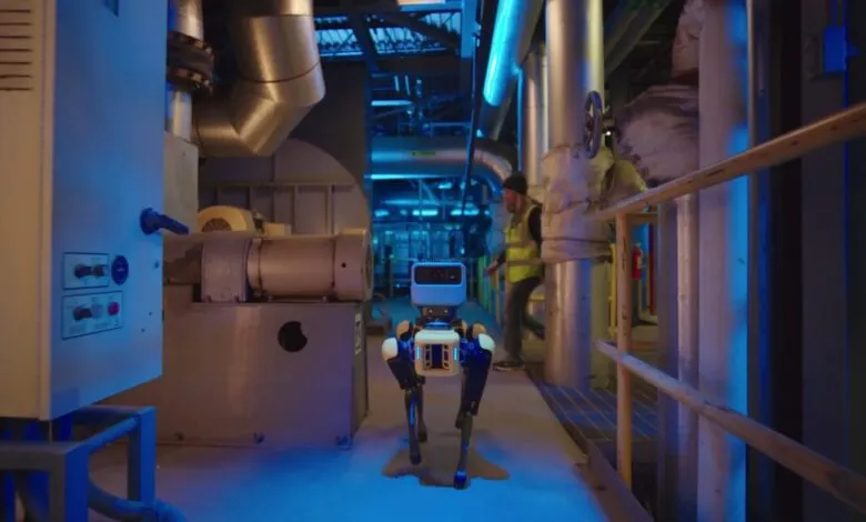 Robot dog walks through industrial setting with pipes and equipment.