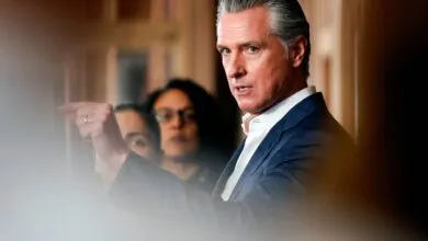 Gavin Newsom points during a press conference, with two women blurred in the background.