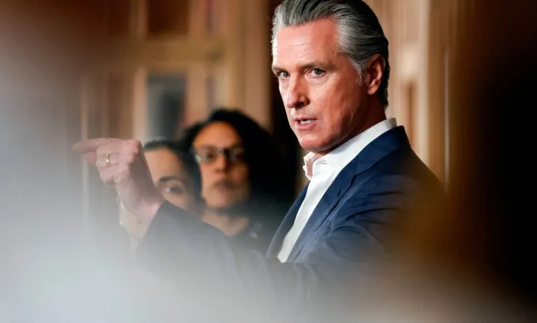Gavin Newsom points during a press conference, with two women blurred in the background.