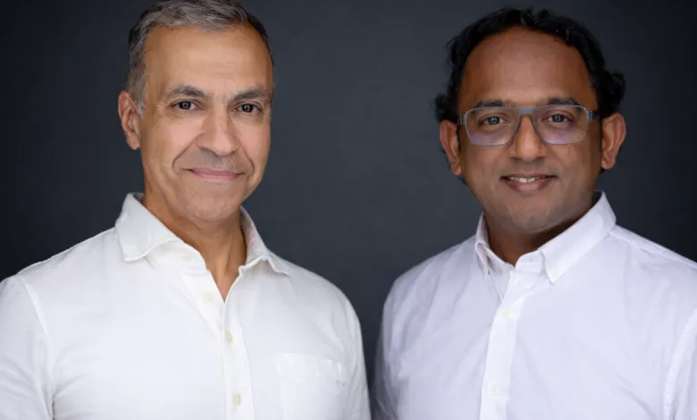 Two men in white shirts pose against a dark gray backdrop, smiling.
