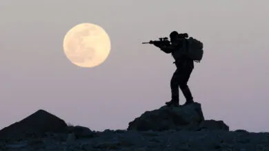 Silhouette of soldier aiming rifle against a full moon.