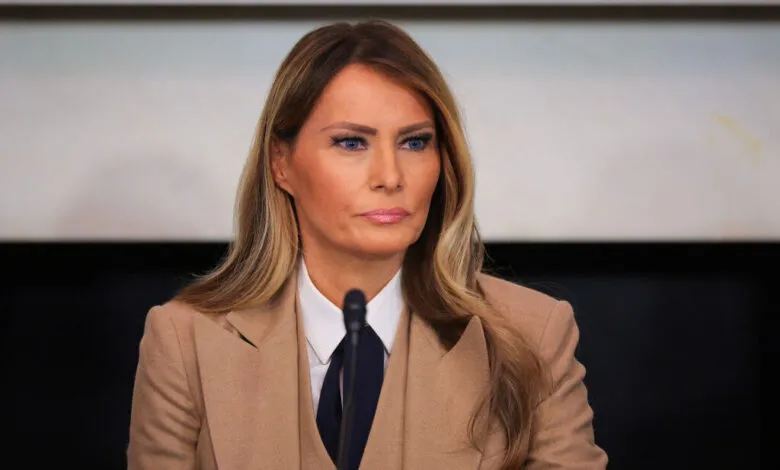 Melania Trump at a podium, wearing a tan suit and blue tie.