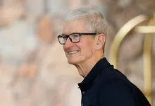 Apple CEO Tim Cook smiles wearing glasses and a dark polo shirt.