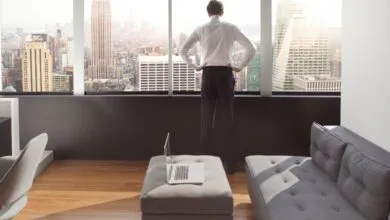 Man in suit looks out window at city skyline, laptop on ottoman.