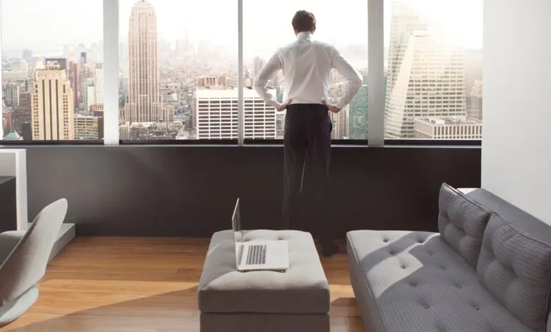 Man in suit looks out window at city skyline, laptop on ottoman.