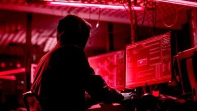 Person in hoodie types at computer screens in a dark, red-lit room.