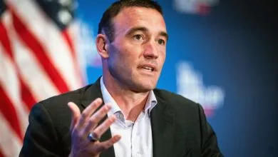 Man in suit gesturing with hand, American flag in background.