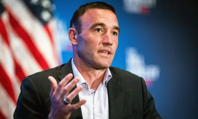 Man in suit gesturing with hand, American flag in background.