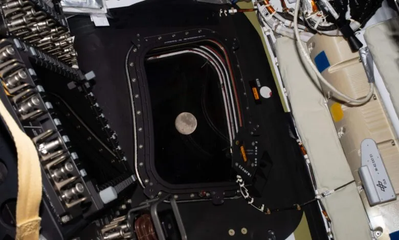 View of the moon through a spacecraft window, surrounded by equipment and wiring.