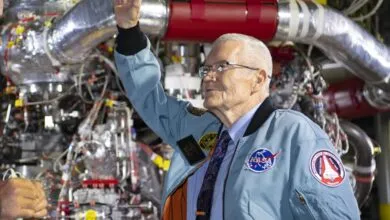 Elderly man in NASA jacket stands before complex machinery, arm raised.