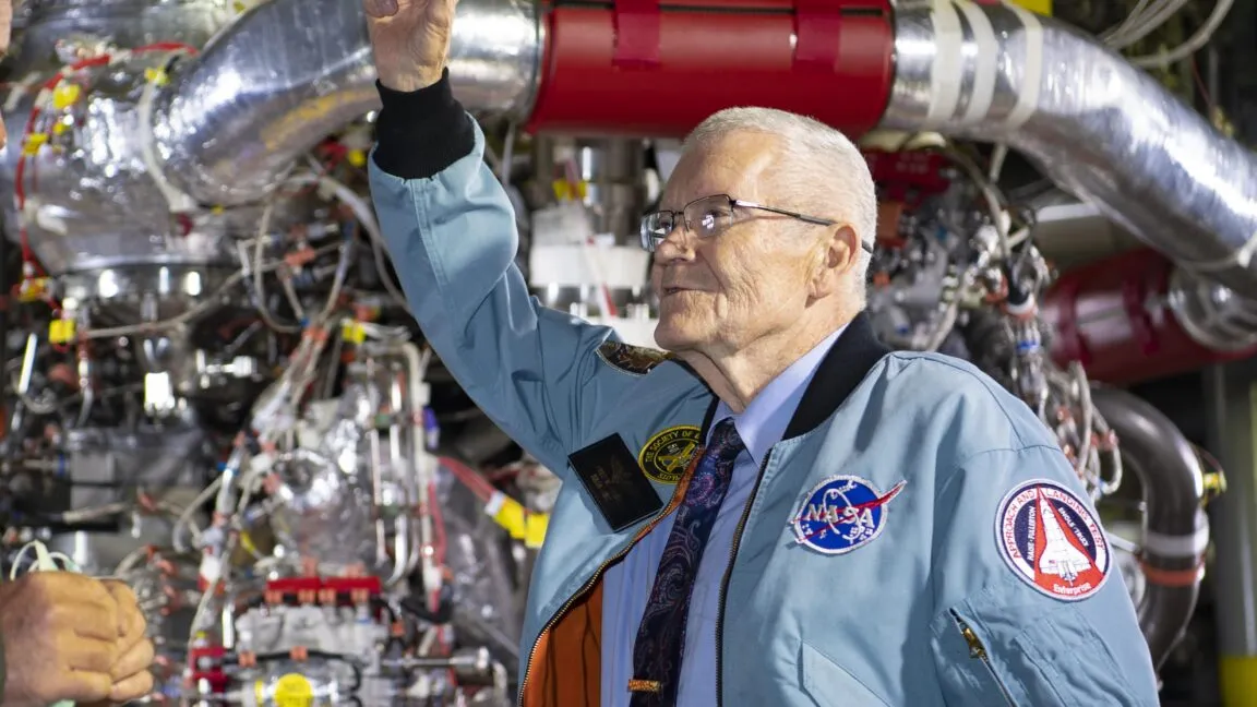 Elderly man in NASA jacket stands before complex machinery, arm raised.