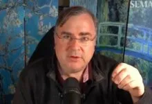 Man with glasses speaking into microphone, art behind him.