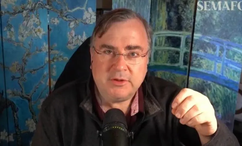 Man with glasses speaking into microphone, art behind him.