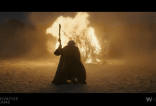 Person in cloak holding staff stands before burning bush in desert landscape.