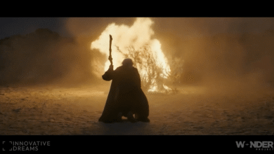 Person in cloak holding staff stands before burning bush in desert landscape.