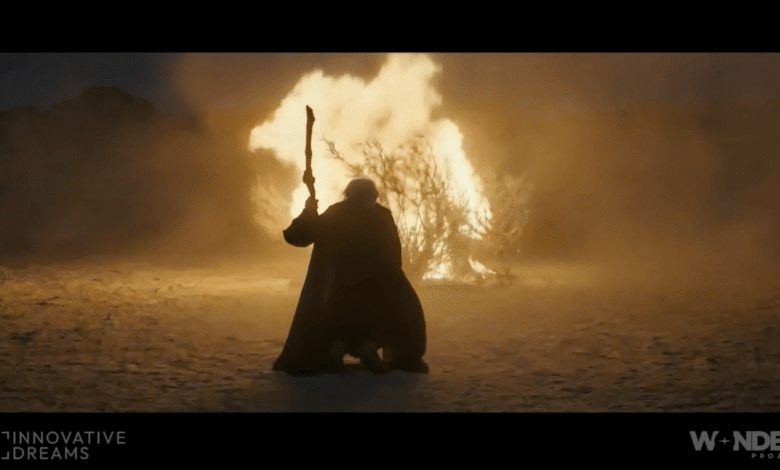 Person in cloak holding staff stands before burning bush in desert landscape.