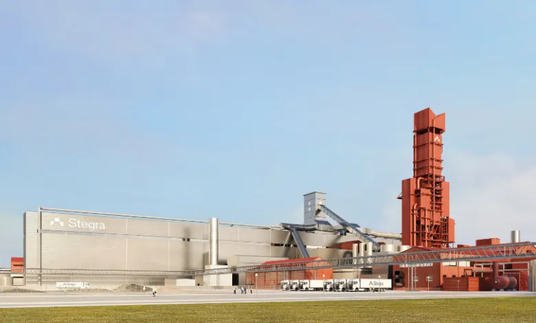 Stegra industrial facility with large building and tall red tower under a blue sky.