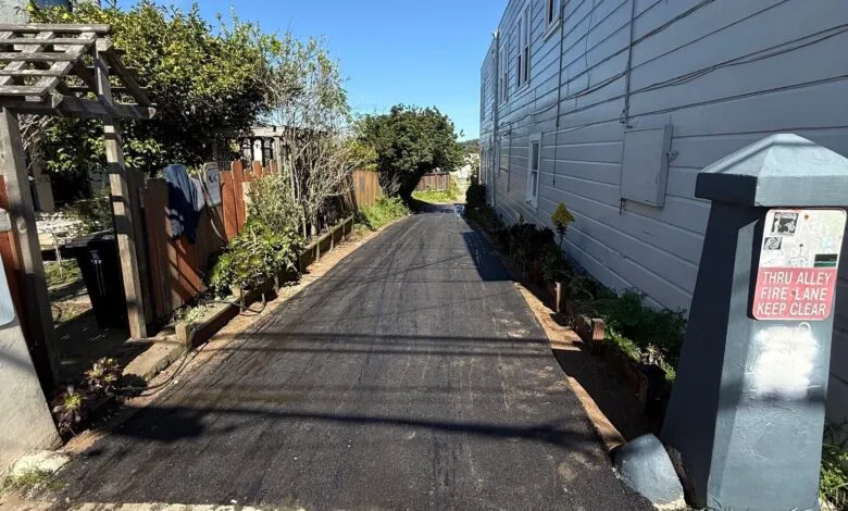 Narrow alleyway between buildings with fresh asphalt and a shadow.