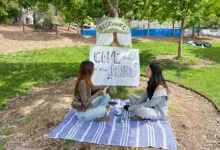 Two young women sit on a blanket in a park, facing each other, with signs promoting connection.