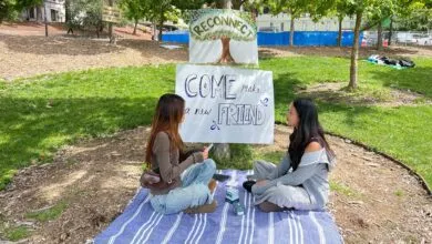 Two young women sit on a blanket in a park, facing each other, with signs promoting connection.