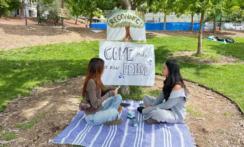 Two young women sit on a blanket in a park, facing each other, with signs promoting connection.