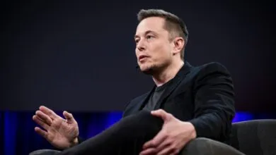 Elon Musk sits in a chair, wearing a black suit and dark shirt.