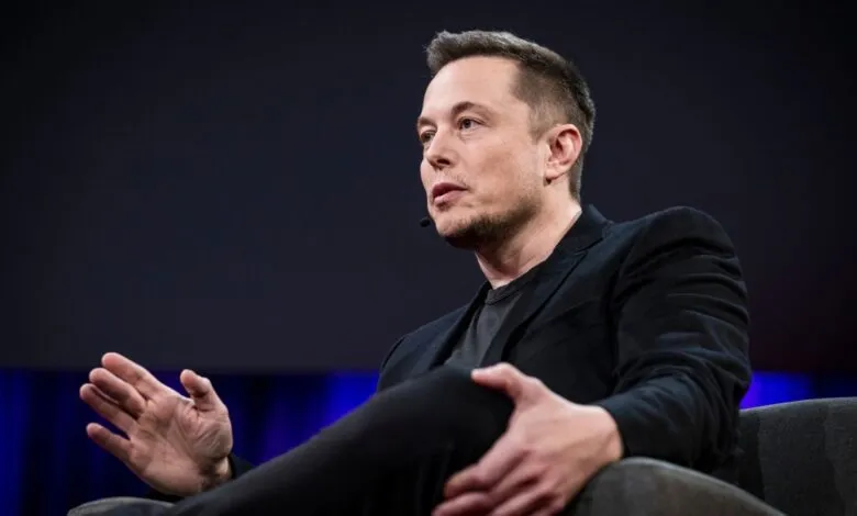 Elon Musk sits in a chair, wearing a black suit and dark shirt.