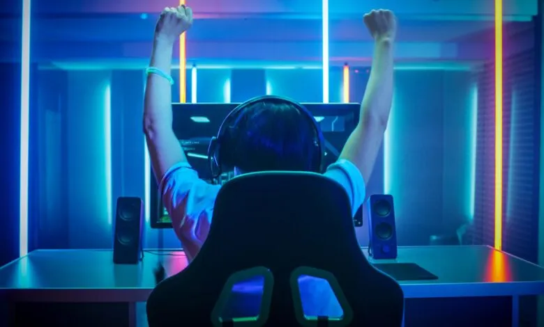 Gamer celebrates victory with arms raised in neon-lit room.