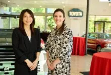 Two women smiling in front of classic cars and a Toyota sign.