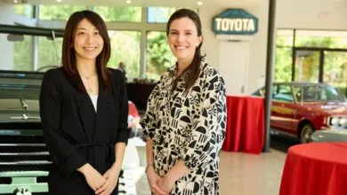Two women smiling in front of classic cars and a Toyota sign.
