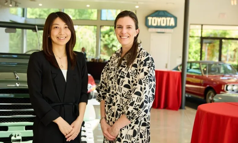 Two women smiling in front of classic cars and a Toyota sign.
