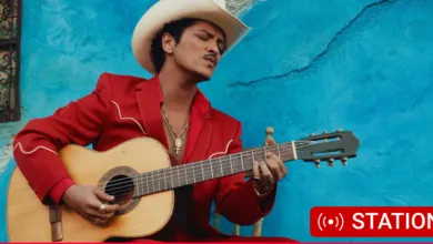 Bruno Mars in cowboy hat plays guitar against a blue wall.