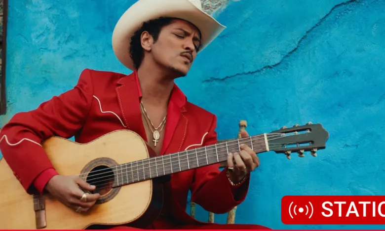 Bruno Mars in cowboy hat plays guitar against a blue wall.