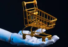 Robot hand holds a miniature yellow shopping cart against a dark background.