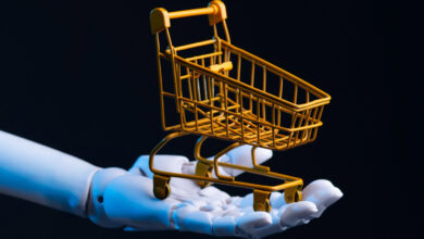 Robot hand holds a miniature yellow shopping cart against a dark background.