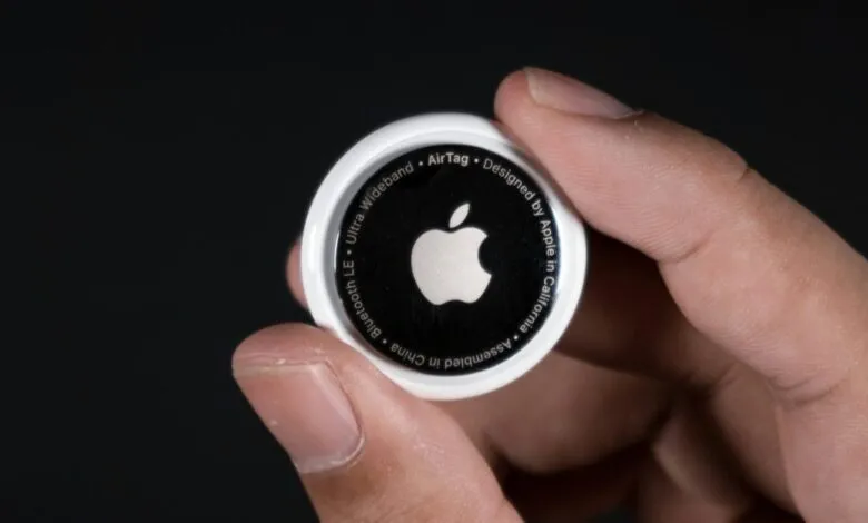 Hand holding an Apple AirTag against a dark background, showing the Apple logo.