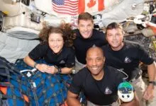 Four astronauts smiling in space station, with US and Canadian flags.